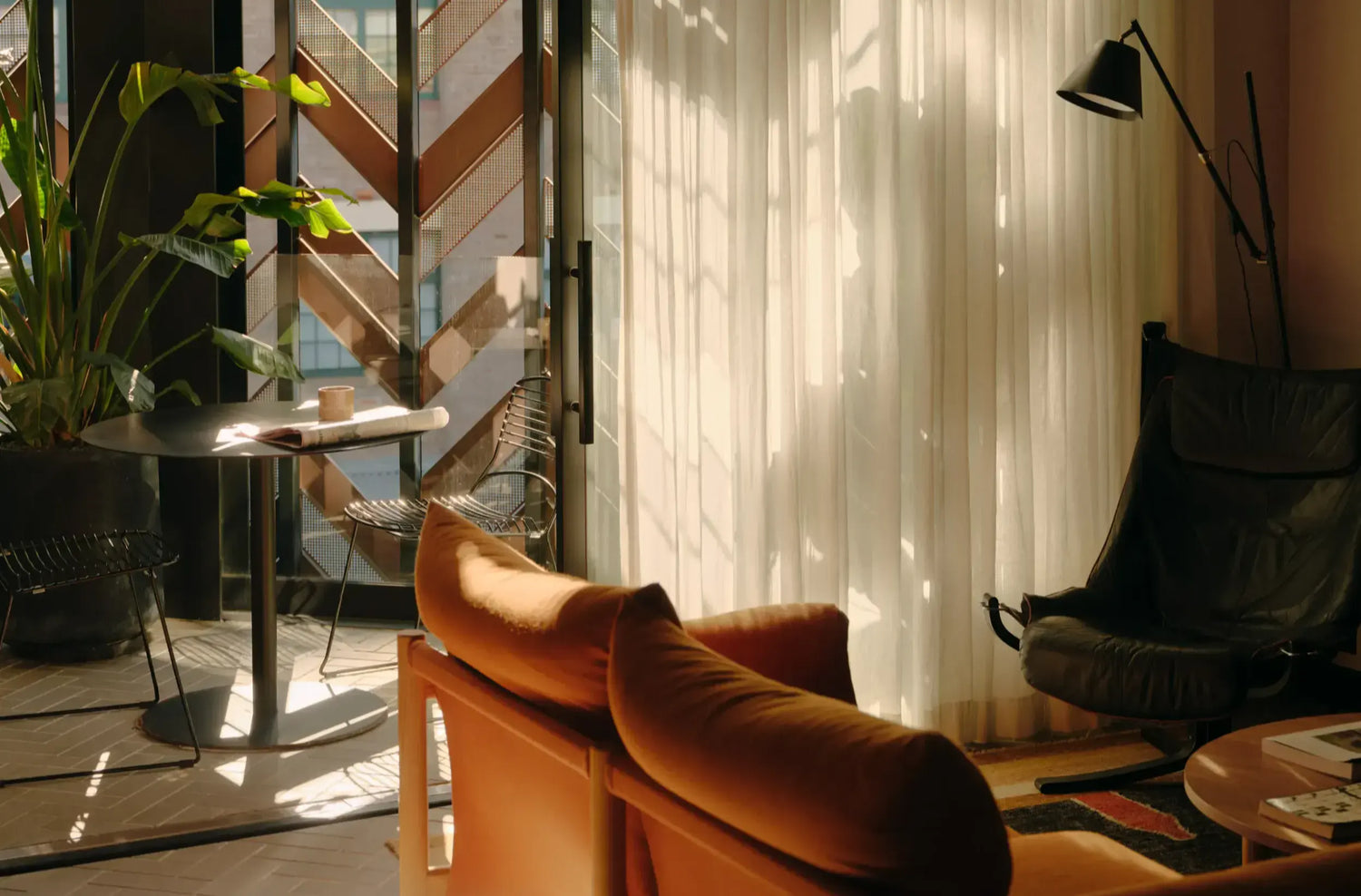 A warm, sunlit modern living room corner with an orange-brown upholstered sofa in the foreground, a black armchair and floor lamp to the right, sheer white curtains, and a glass door opening to a balcony or patio with plants and outdoor light.