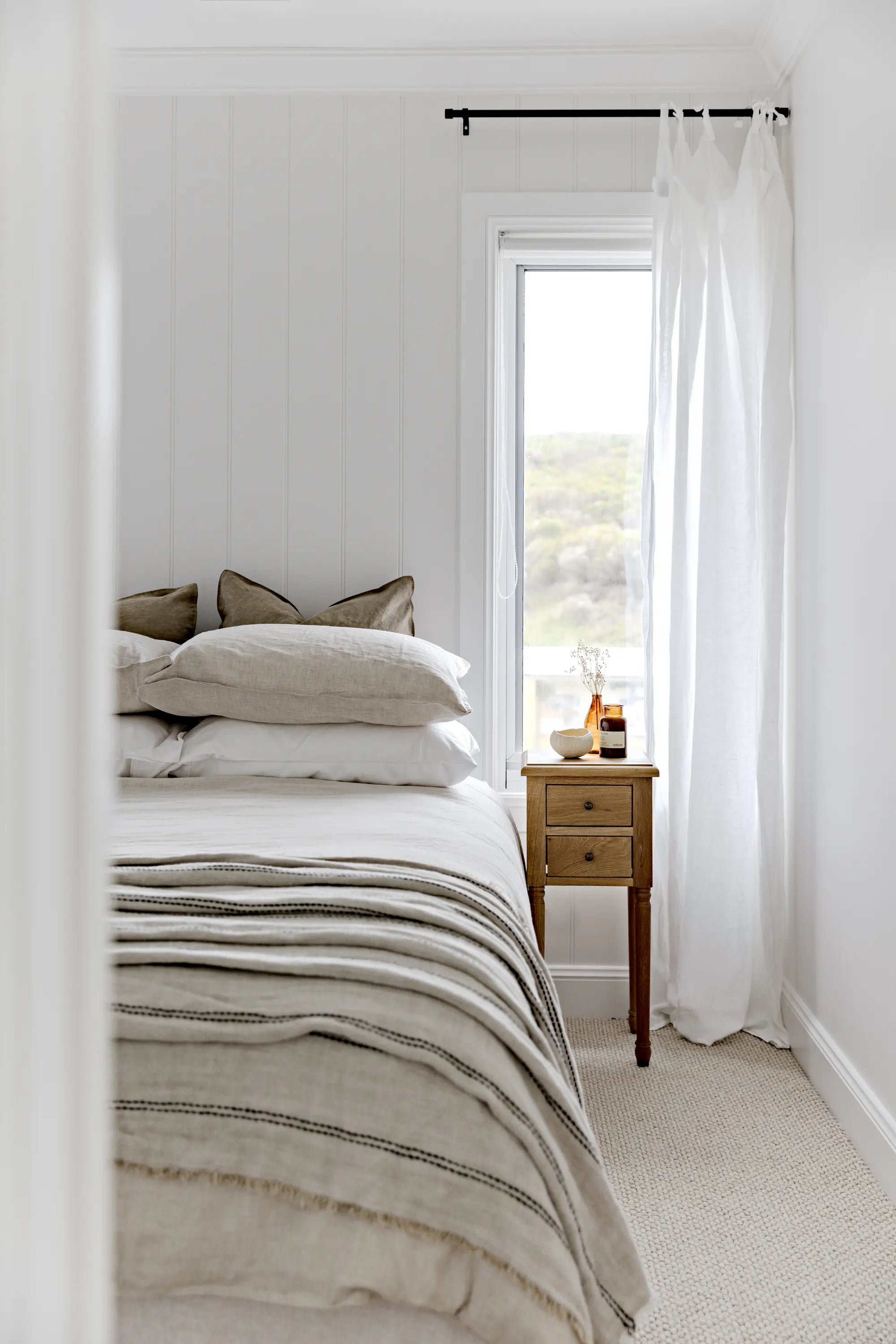 A styled bedroom interior featuring a bed dressed in neutral beige and white linens, a small wooden nightstand with bottles, and a tall window with white sheer curtains and a view of an outdoor landscape.