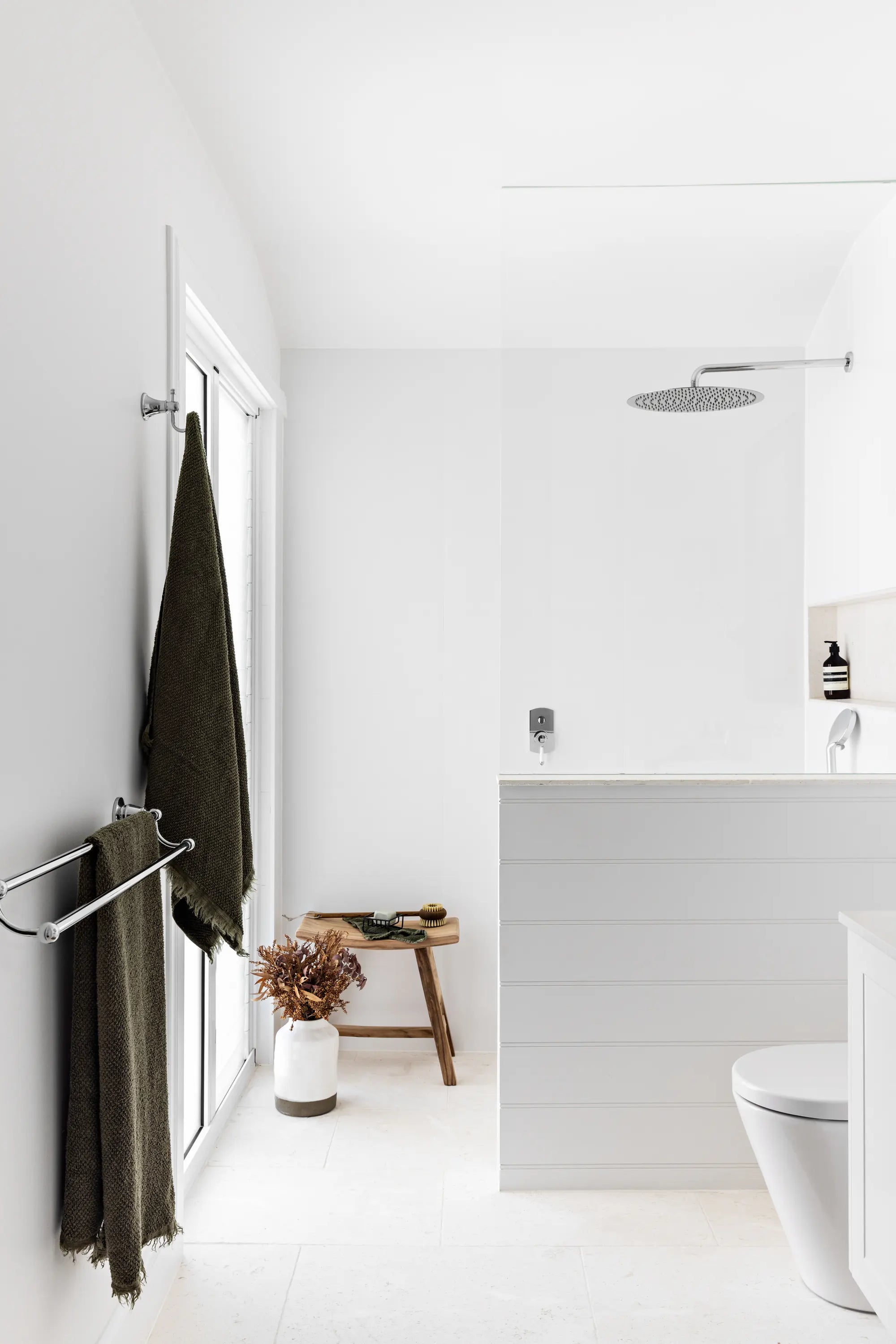 A modern minimalist bathroom with white walls and fixtures, featuring a dark olive green towel hanging on a wall-mounted rack, a small wooden stool with dried flowers, and a white toilet and vanity.