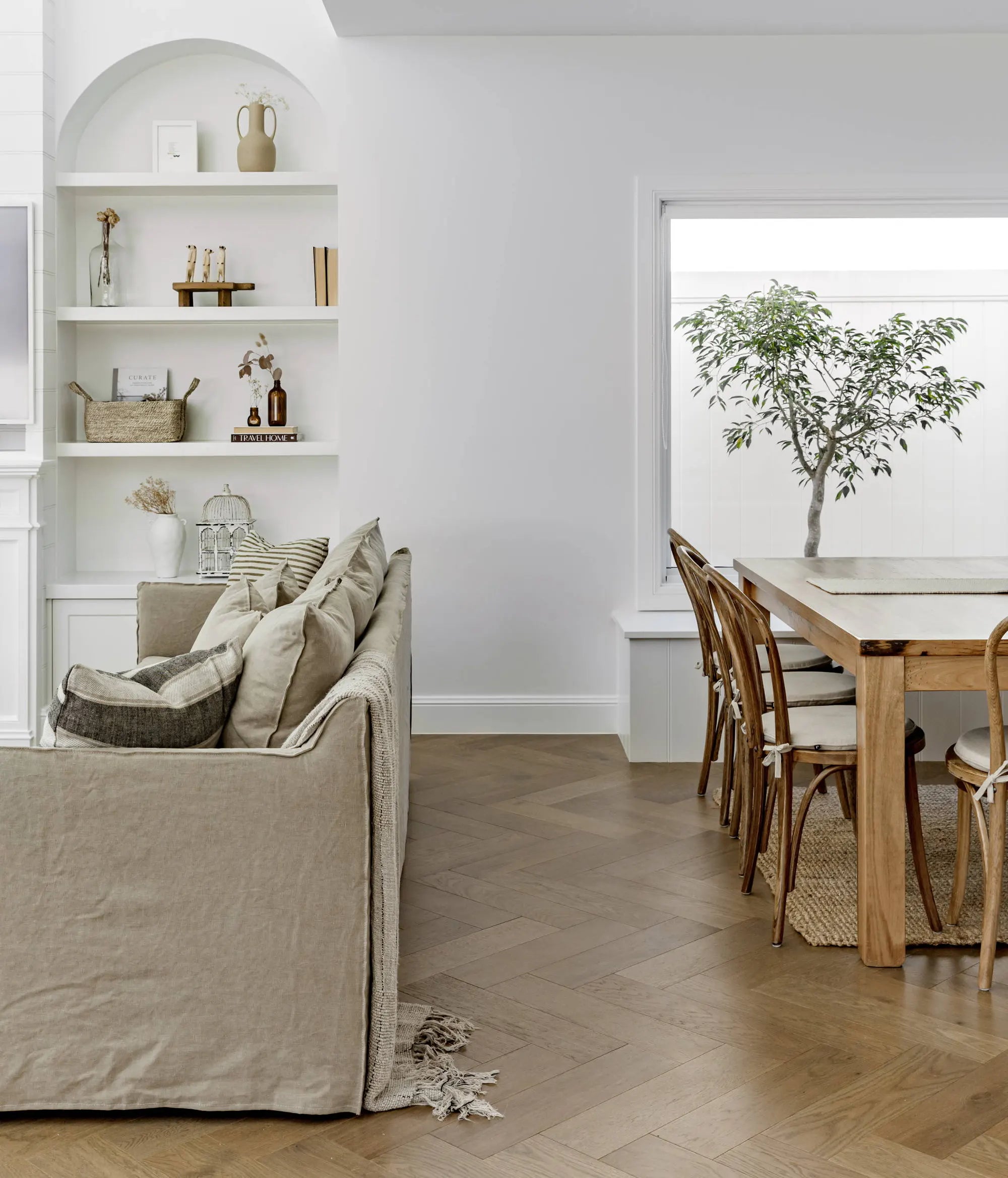 A styled interior scene showing a beige slipcovered sofa with pillows in a bright room, next to a wooden dining table and chairs, with built-in white shelves and a window view of a tree.