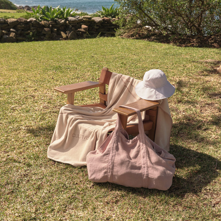 A Beach Towel in Rose on a lawn chair, featuring a Sol Linen Hat and Frankie Bag in Dusk
