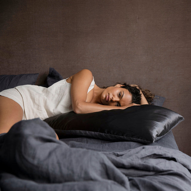 A woman relaxes on a bed dressed in Slate bedlinen, and a Silk Flip Pillowcase in Slate. Size: Single Standard, Set of 2 Standard