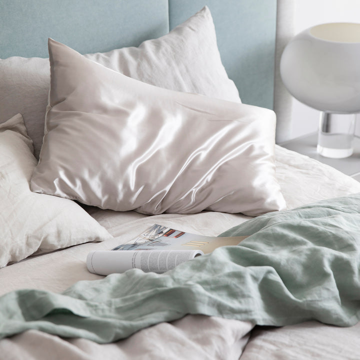 Bed styled with the Smoke Grey Duvet Cover set, a Smoke Grey Fitted sheet, Sage Flat Sheet with Border and a pair of Smoke Grey Silk Linen Flip Pillowcases. Also featured is an open magazine positioned on top of the sheets.