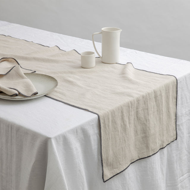 Cara Edged Table Runner in Slate on a Linen Tablecloth in White. Size: 50cm x 200cm, 50cm x 300cm