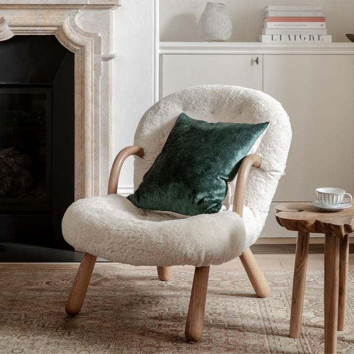 A Talik Velvet Cushion in Pine, styled with an armchair and a side table in front of a fireplace. Size: 50 x 50cm, 60 x 40cm