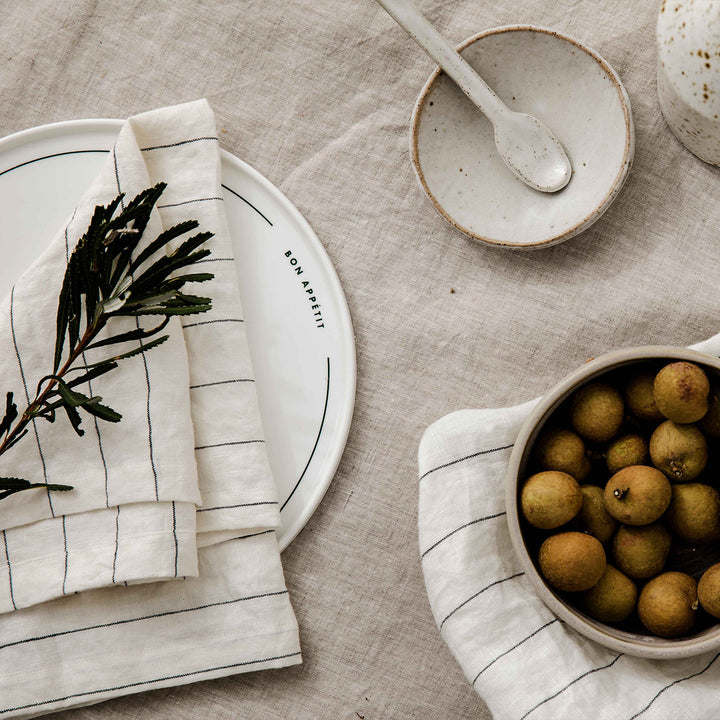 A table setting featuring the Linen Tablecloth in Natural, Linen Table Napkins in Pencil Stripe, a ceramic bowl, plate and spoon, and longan fruit