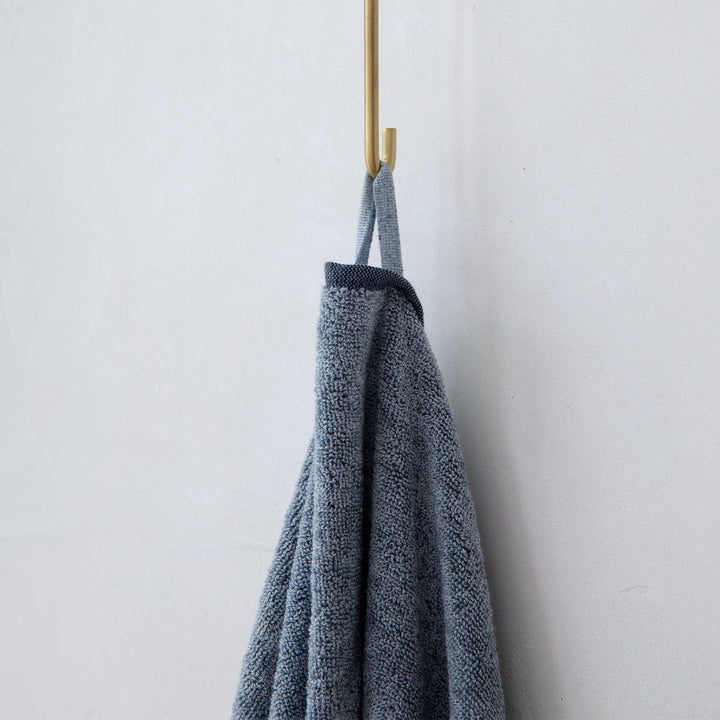 A close up of a denim towel hanging on a hook. Sizes: Bath Towel Bundle - 28" x 55", Bath Sheet Bundle - 35" x 69"