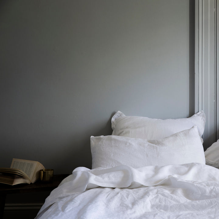 A bed dressed in White bedlinen styled with a bedside table, against a grey wall.